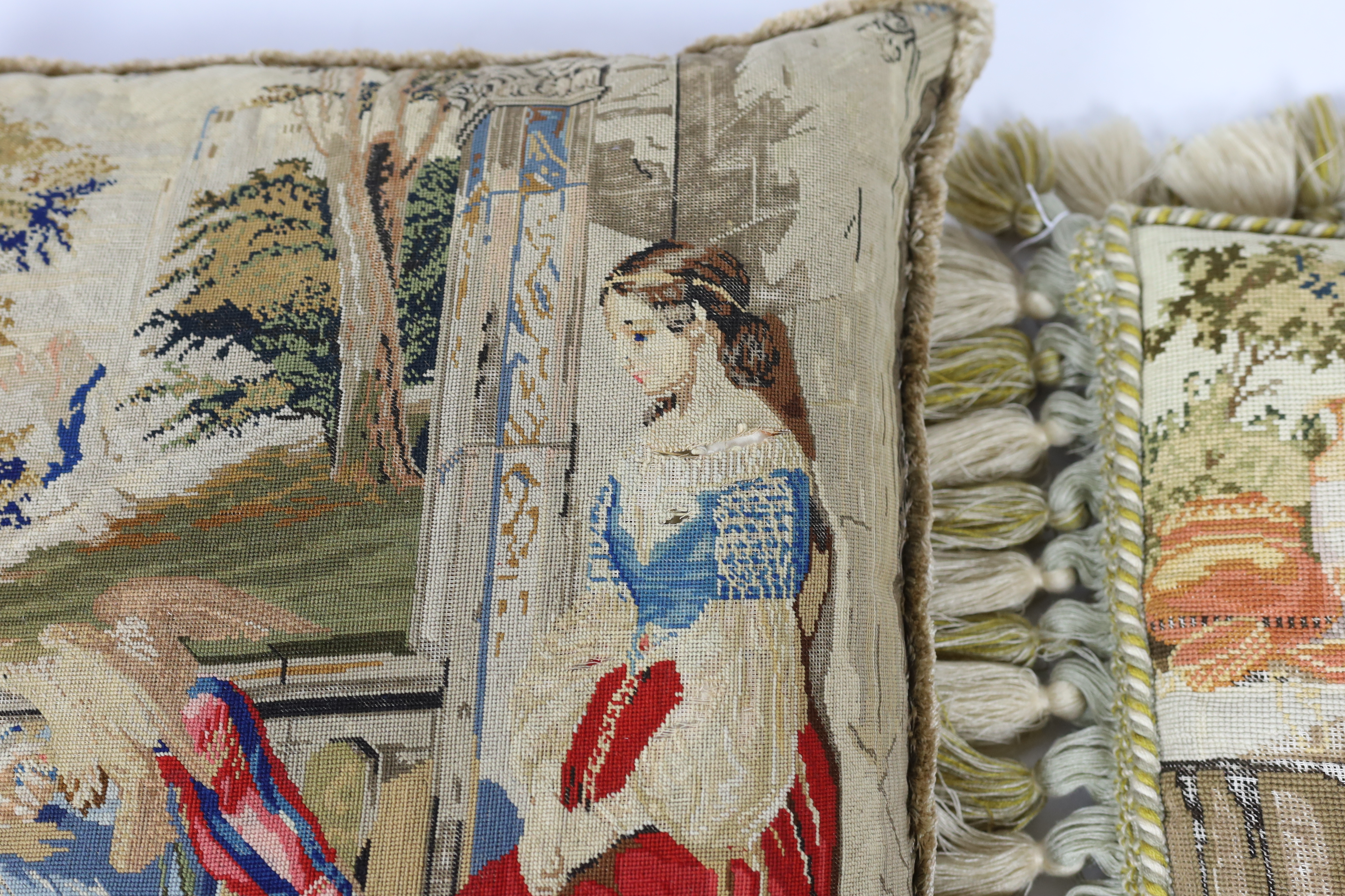 Three Victorian Berlin wool worked cushions possibly of classical biblical scenes, all three cushions edged with braiding and fringing, largest cushion 61cm wide, 65cm high